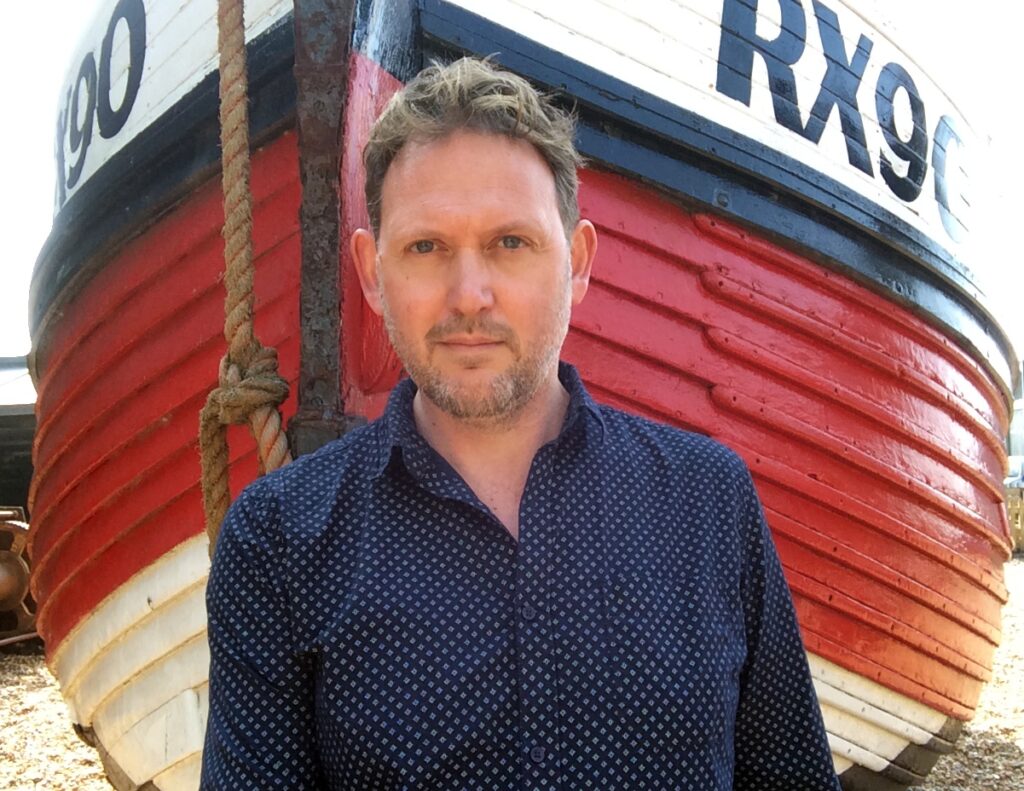 Bir adam, arkasındaki renkli balıkçı botuyla birlikte duruyor.