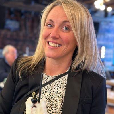 Bolton'da gülümseyen bir kadın, içecek tutuyor ve şık bir ceket giymekte.